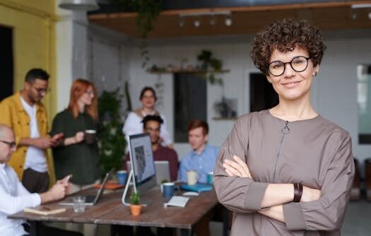 Smiling businesswoman with curly hair stands confidently in a modern office space with colleagues.